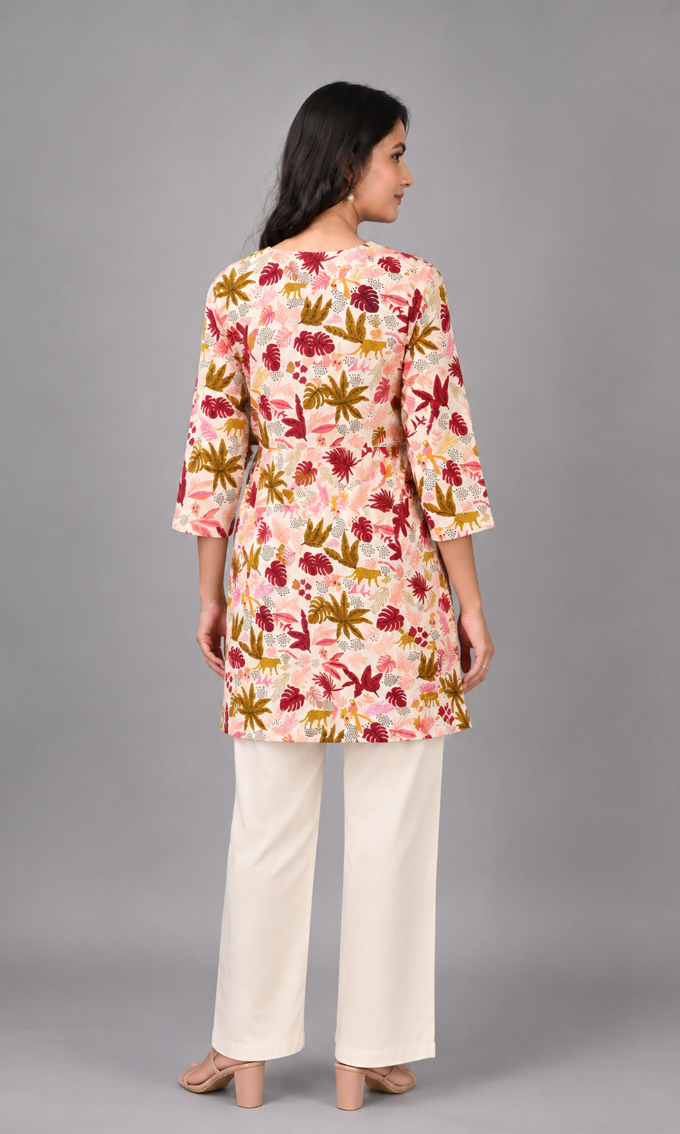 A woman shown from the back wearing a light pink floral kurta with three-quarter sleeves and cream wide-leg pants, set against a solid gray studio background.