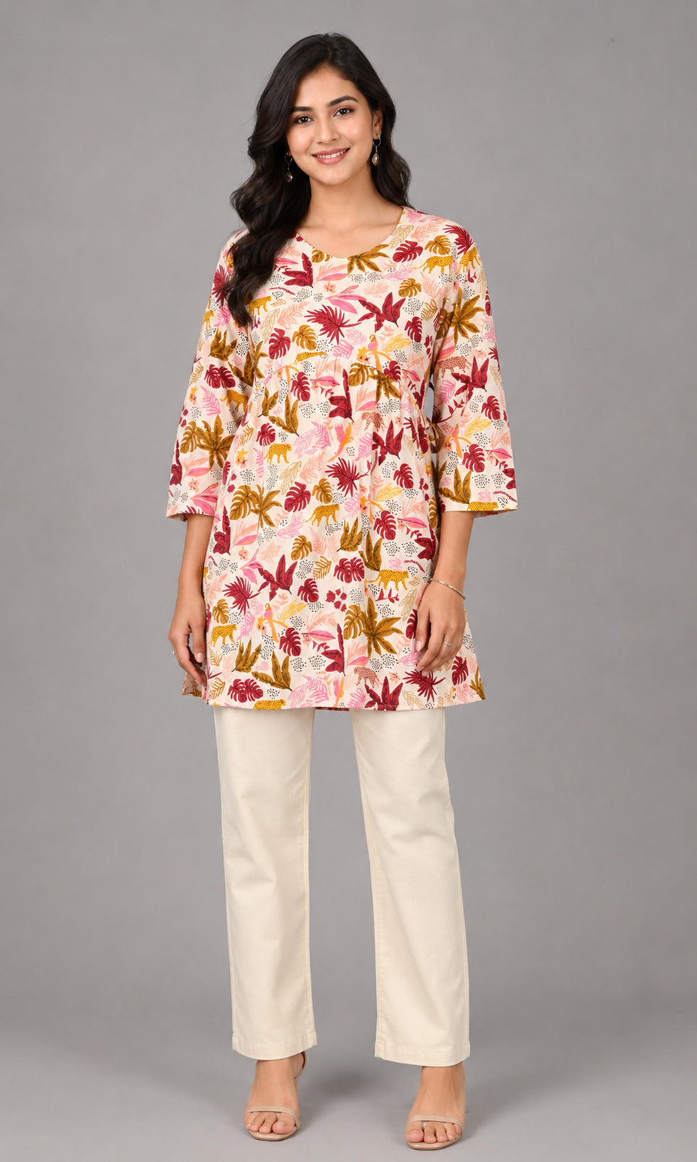A woman modeling a three-quarter sleeve kurta with a pink, red, and gold floral print, paired with cream-colored straight-leg trousers, against a solid gray studio background.