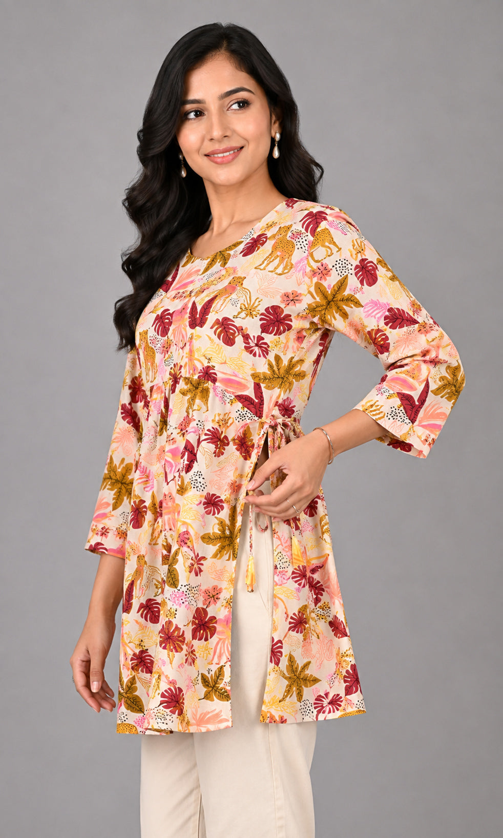A woman modeling a peach-colored three-quarter sleeve tunic top with a multicolor floral and animal print, paired with cream pants, against a solid gray studio background.